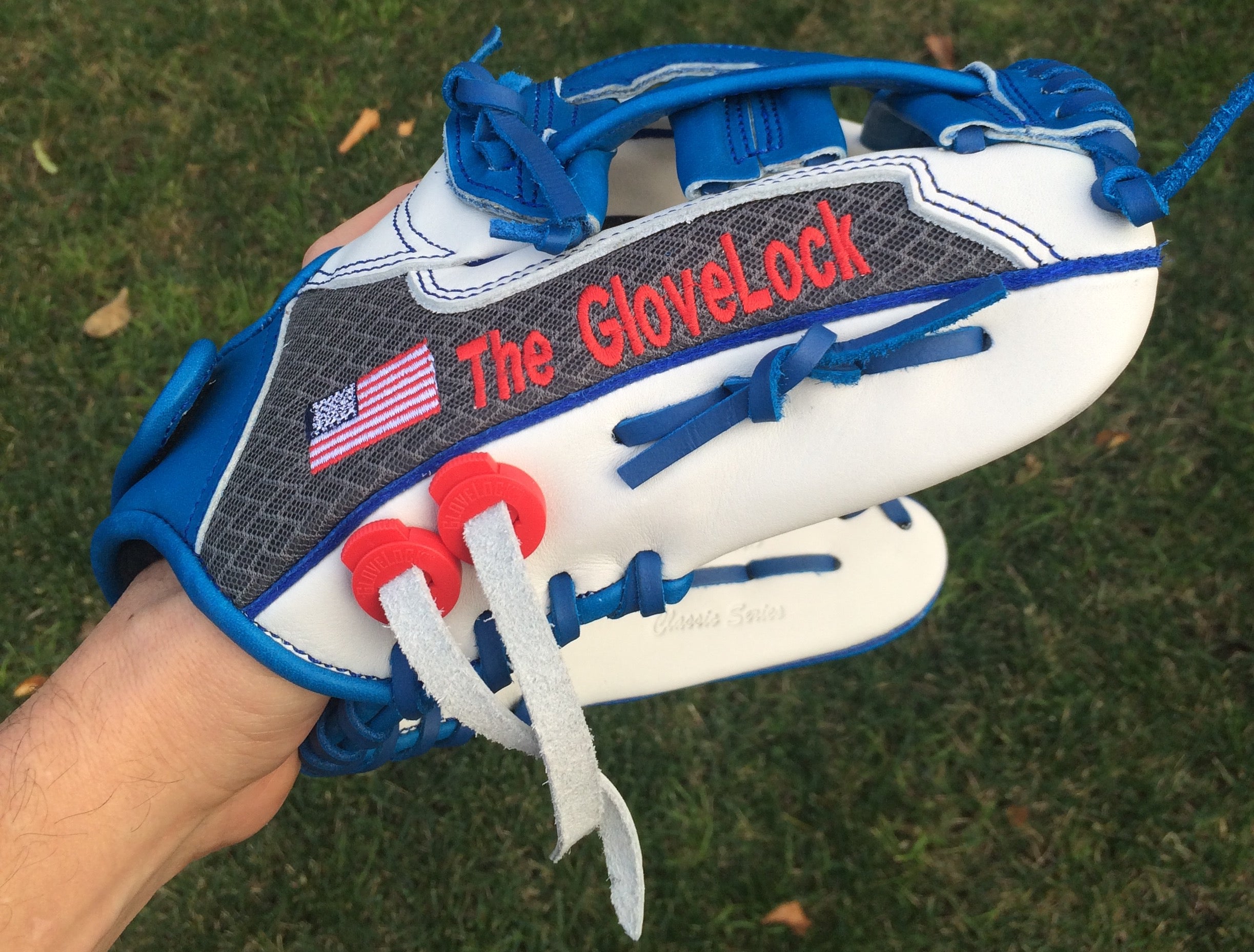 A hand holding a white and blue baseball glove with 'The GloveLock' embroidered in red on the back, set against a background of green grass.